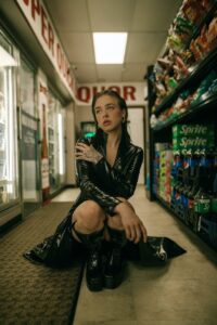 young woman in black pvc matrix style trench coat sitting on the floor at a supermarket aisle